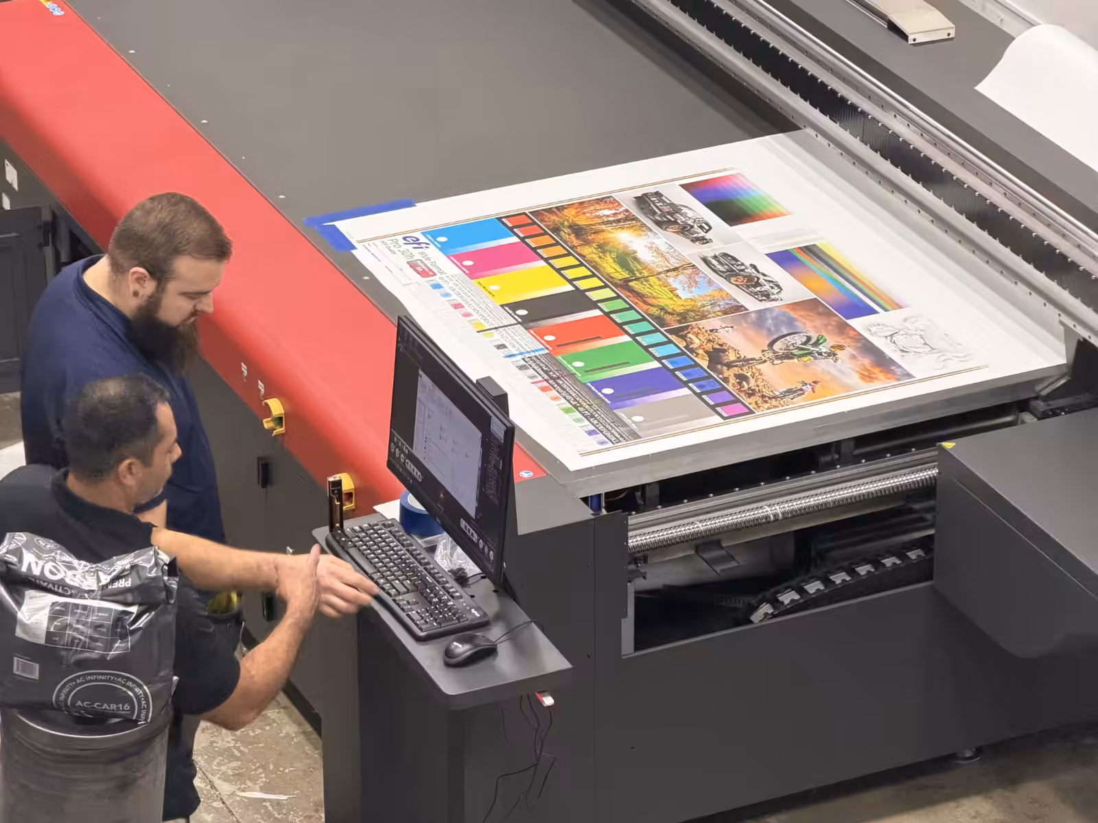 Two men operate a large industrial printer, reviewing colorful printed wraps and test sheets on the printer bed while checking settings on a nearby computer.