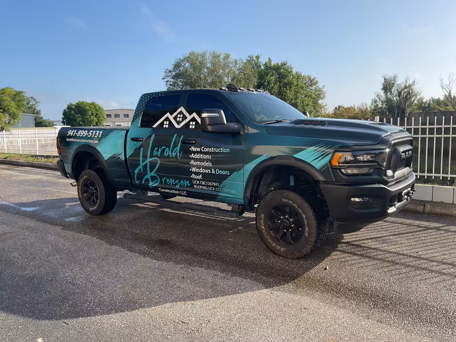 A black and teal pickup truck with World Construction LLC branding and custom wraps is parked on pavement, displaying a phone number and construction services. Trees and a white fence are in the background.