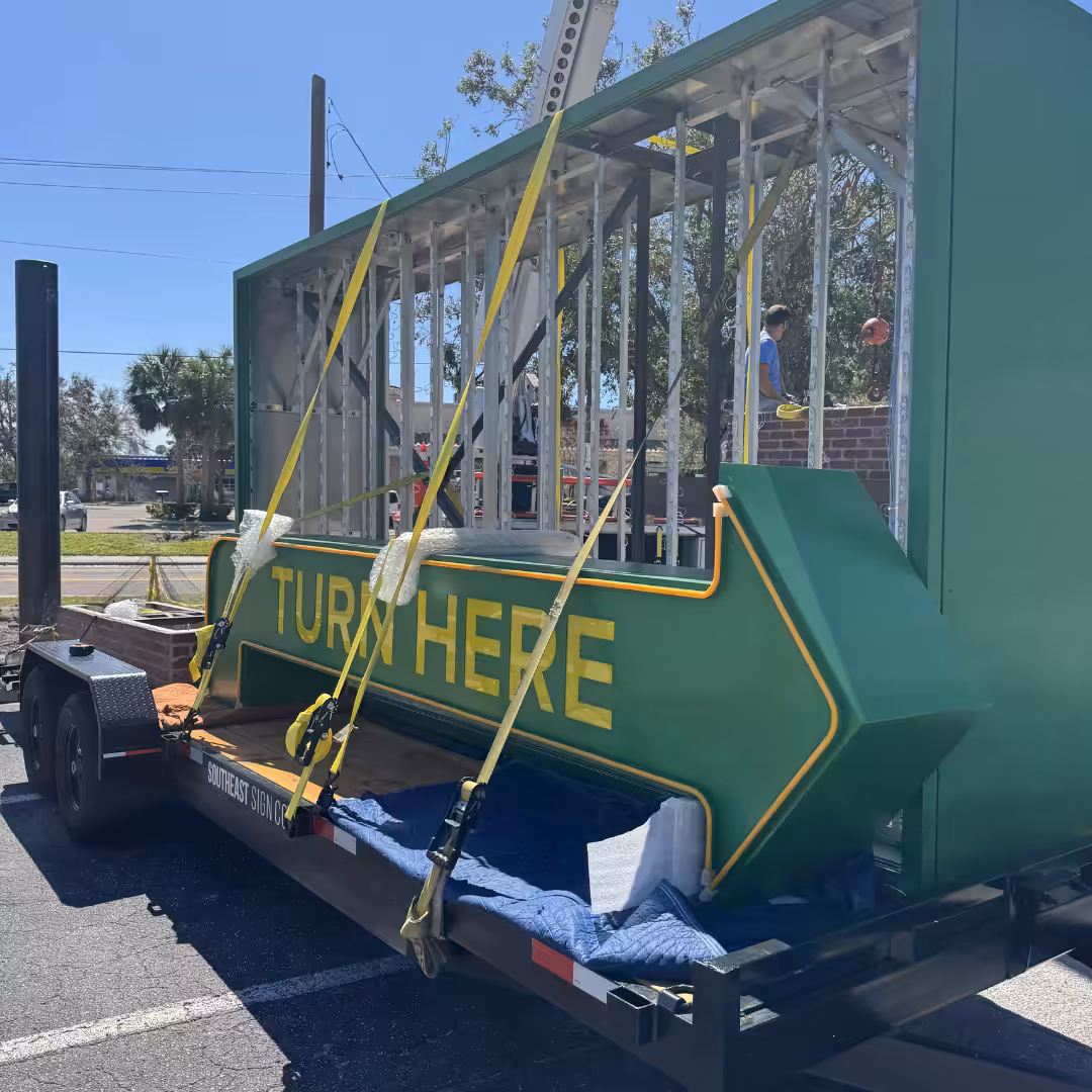 Large green arrow sign with yellow text TURN HERE strapped to a trailer outdoors, serving as directional storefront signage. People are visible in the background near a construction frame.