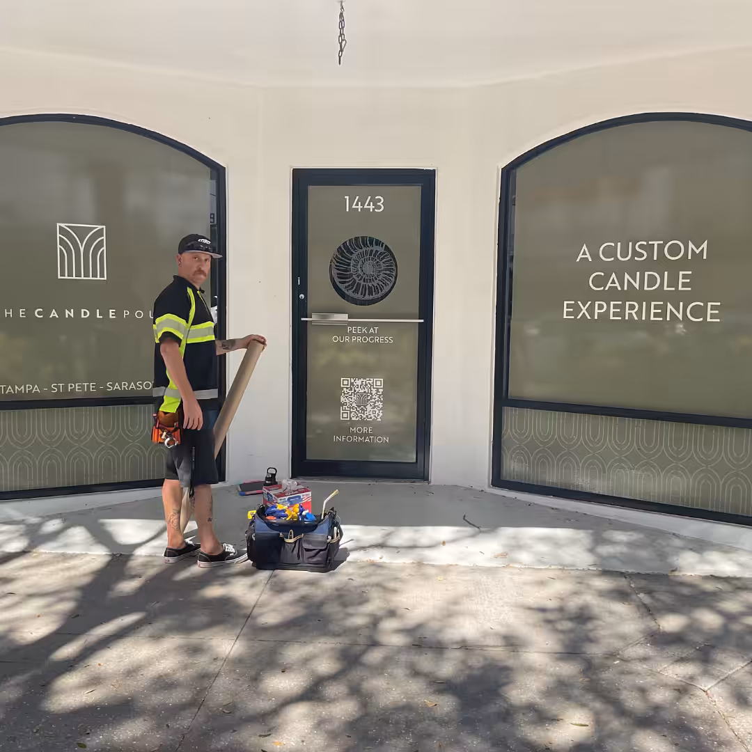 A worker in a safety vest stands with tools and supplies outside a storefront displaying window graphics and signs reading A Custom Candle Experience and The Candle Pour at 1443.