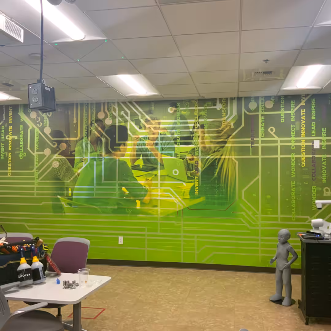 A mural of students working with technology is on a green wall with circuit patterns in a classroom. Indoor business signs add detail, while a mannequin stands nearby; tables and tools are visible in the foreground.