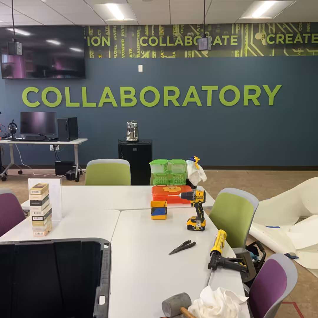 A workspace labeled COLLABORATORY features tables, chairs, tools, boxes, electronic equipment, and striking indoor business signs displaying words like INNOVATION and CREATE above the main sign.