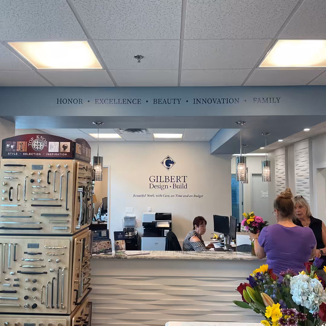 Reception area of a design business with employees at the desk, floral arrangements in the foreground, indoor business signs, and cabinet hardware displays. Words like HONOR and FAMILY are written on the wall above the desk.
