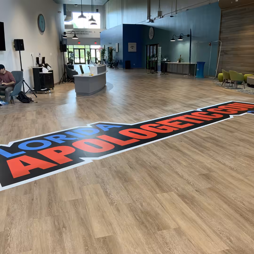 A large indoor business sign on a wood floor reads FLORIDA APOLOGETICS, partially cut off. The spacious room features a reception desk, green chairs, and people setting up equipment.