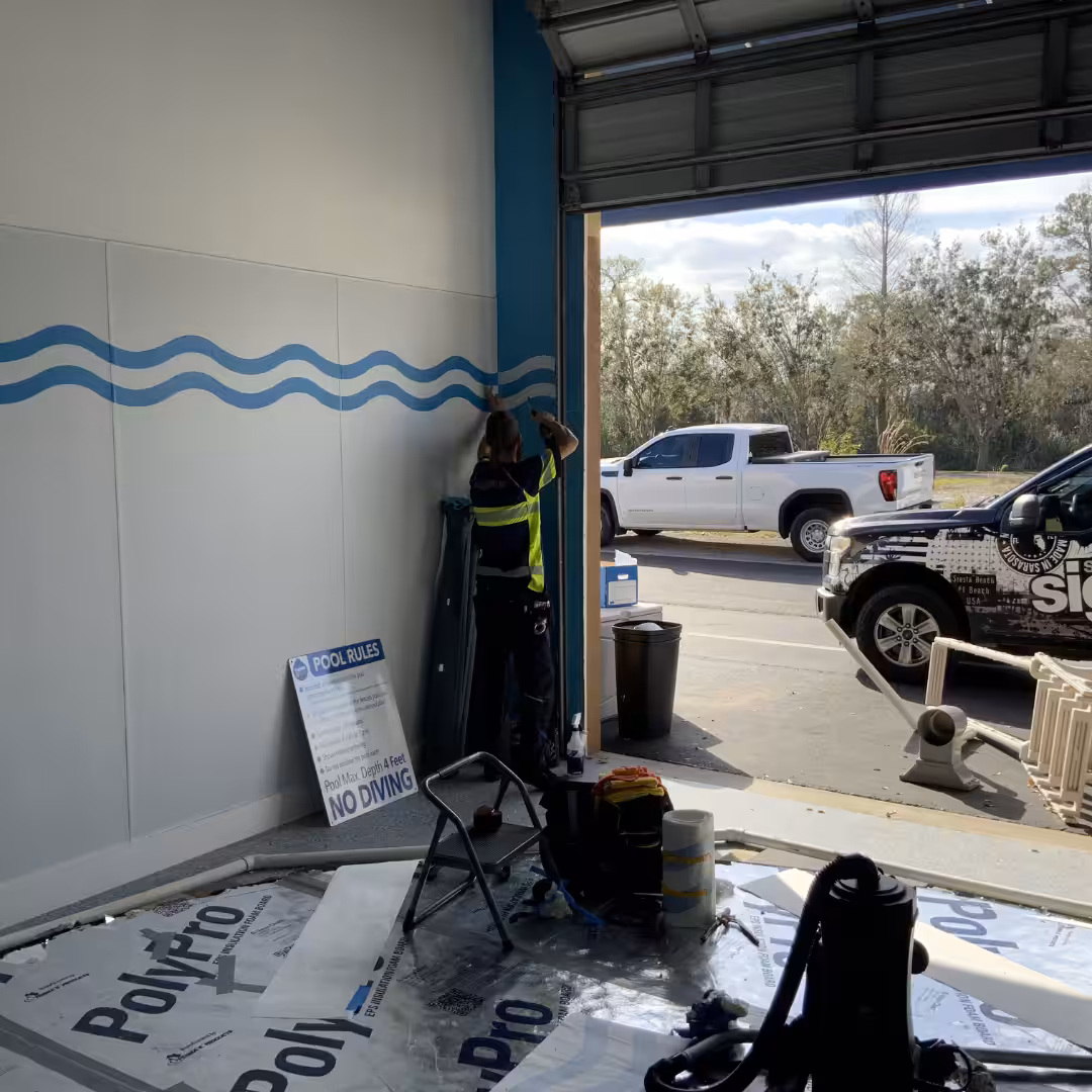 A person in a safety vest installs or repairs electrical wiring inside a garage with white walls, where indoor business signs are stored among various tools. Two trucks are parked outside in daylight.