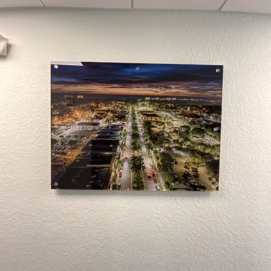 A framed aerial photograph of a city street at dusk hangs on a textured white wall, its illuminated buildings echoing the glow of indoor business signs and parked cars beneath a darkening sky.