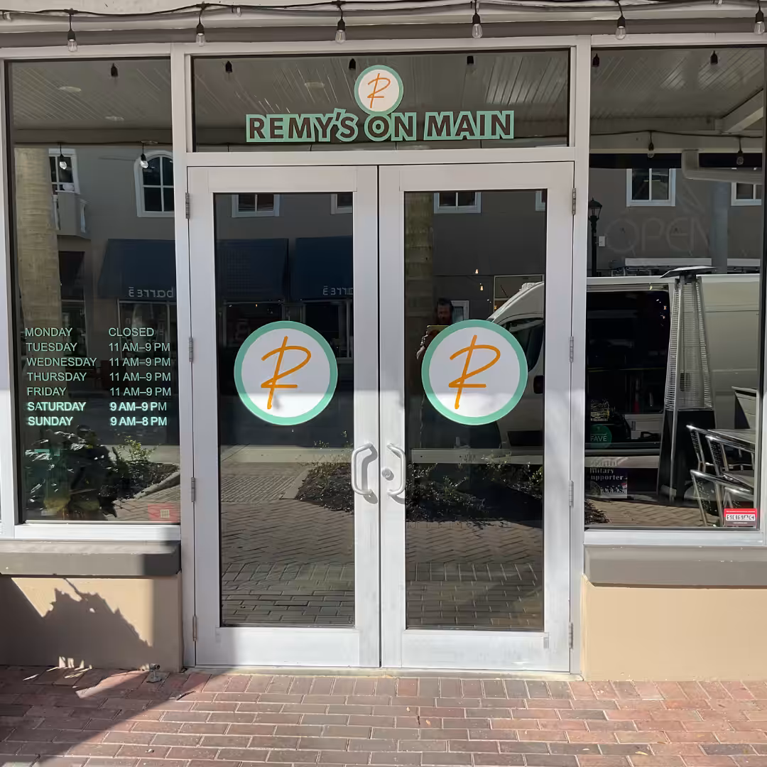Glass doors of Remys On Main restaurant feature business hours posted and window graphics with two circular logos, each sporting an orange R. The sunlit entrance opens onto brick pavement outside.