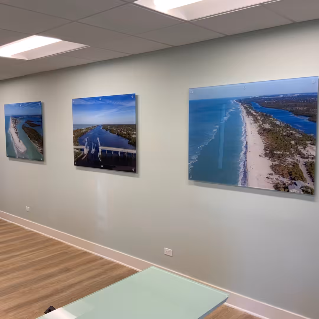 Three large aerial photos of a coastal beach and bridge are displayed on a pale green office wall above light wood floors and a white table, creating an atmosphere as inviting as stylish indoor business signs.