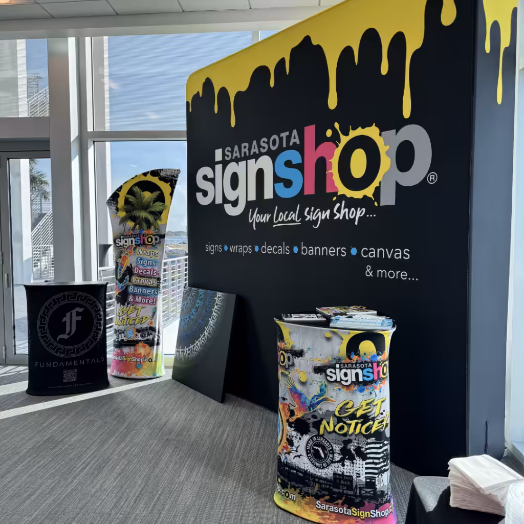 Indoor display for Sarasota Sign Shop, featuring a large sign with dripping yellow paint, colorful promotional stand, banners, and eye-catching tradeshow graphics for signs, wraps, decals, canvas, and more.