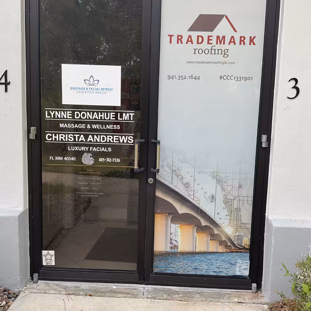 Glass door with window graphics for Trademark Roofing, Massage & Facial Retreat, and luxury facials. A bridge photo is on the right, contact info and license numbers are listed, and building numbers 4 and 3 are visible.