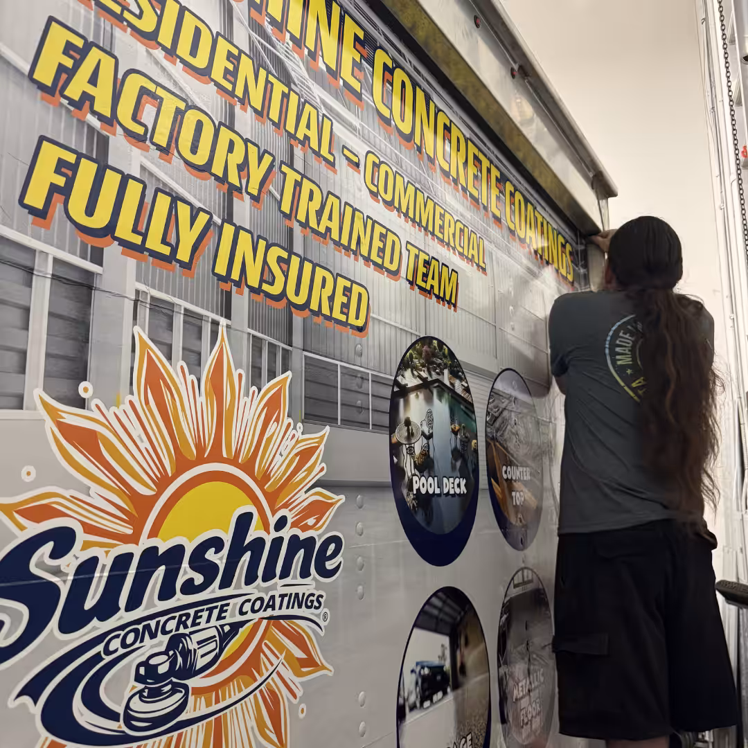 A person with long hair applies a vinyl wrap to a truck and trailers displaying Sunshine Concrete Coatings—Residential, Commercial, Fully Insured—Factory Trained Team, with images of finished work.