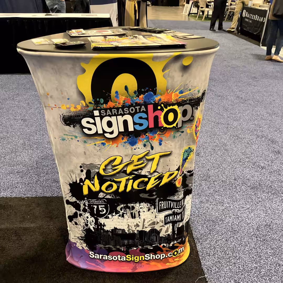 A colorful promotional booth for Sarasota Sign Shop displays Get Noticed! with Tradeshow Graphics, paint splatters, contact info, and brochures on top, at a trade show with blue carpet.