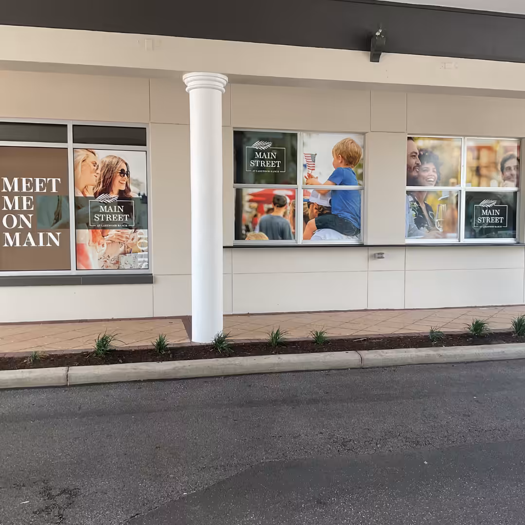 Storefront with large windows featuring window graphics of people smiling, eating, and drinking. The text MEET ME ON MAIN and MAIN STREET appears on the windows. A white pillar and small plants line the sidewalk.