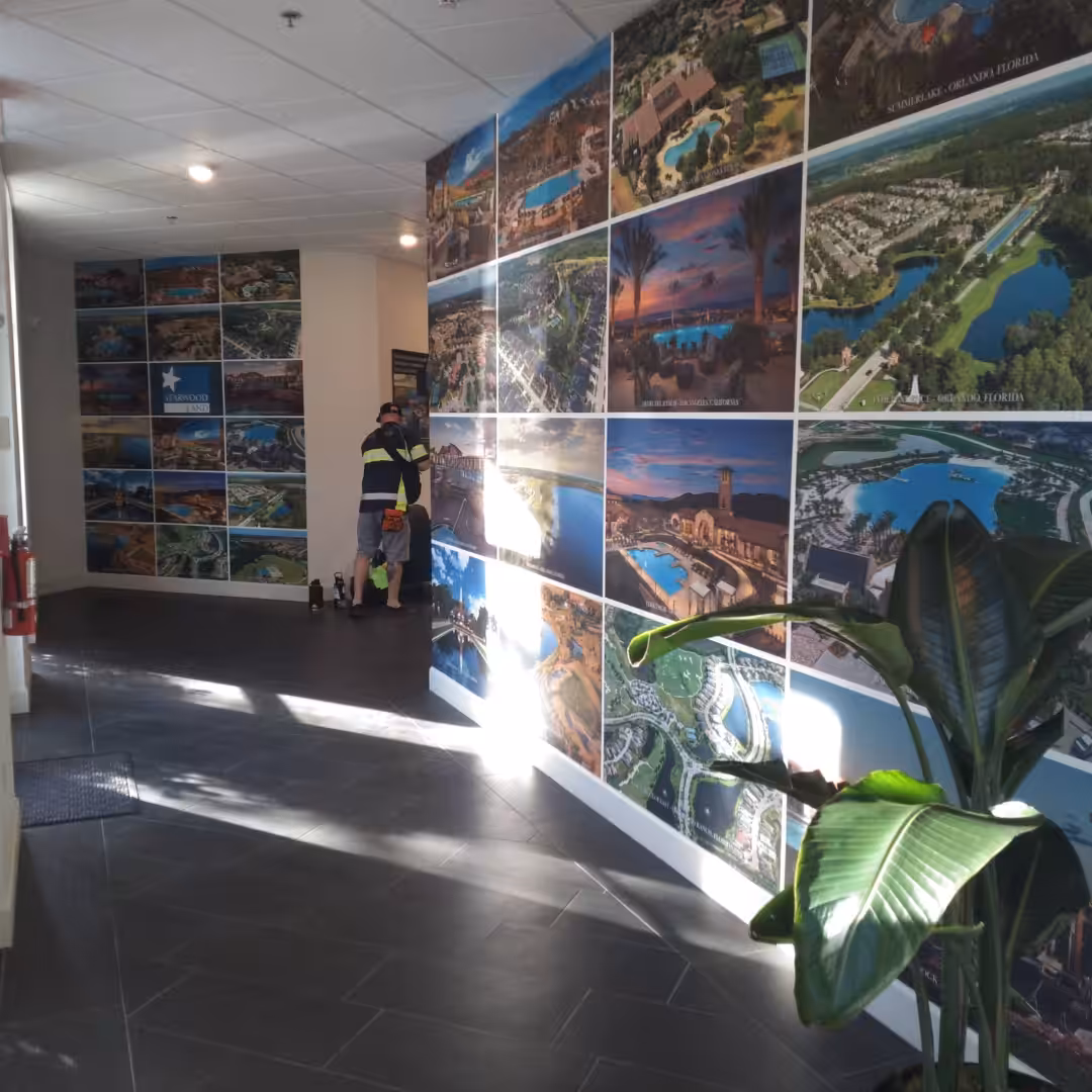 A hallway with tiled floors, indoor business signs, a large plant, and walls decorated with a collage of aerial landscape photographs. Two people in reflective clothing stand near the end of the hallway.