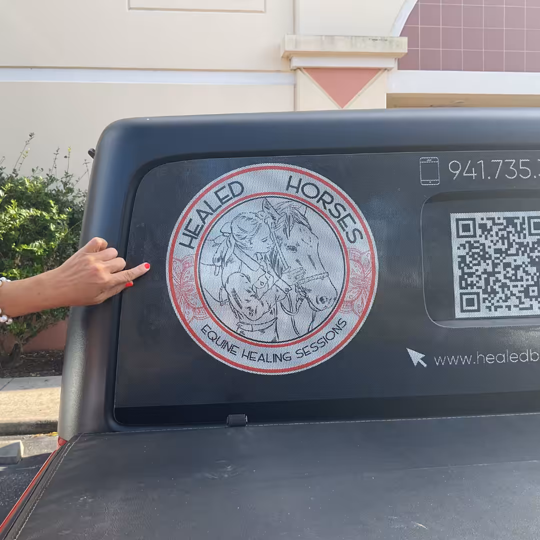 A person points to a circular Healed Horses Equine Healing Sessions decal with horse drawings and Window Graphics on a vehicle window. A QR code and partial phone number are also visible.