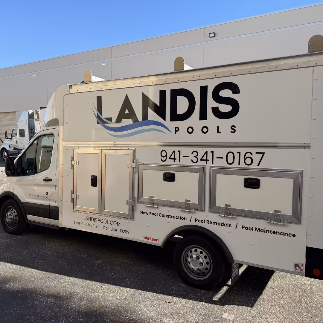 A white Landis Pools service van and trailers are parked outside a building, displaying the company’s logo, phone number, and services like new pools, remodels, and maintenance.