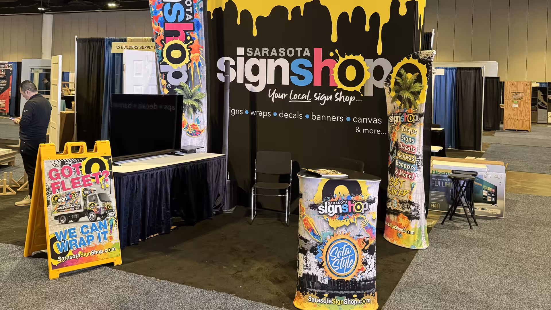 A colorful Sarasota Sign Shop booth showcases Tradeshow Graphics, including banners, a yellow sandwich board, a branded table, and a wrapped column against a black-and-yellow dripping paint backdrop.